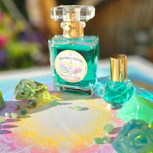 Puede incluir: Dos botellas de l&iacute;quido verde con tapas y etiquetas doradas. La botella m&aacute;s grande tiene una etiqueta que dice "Andara Crystal Magical Aura Spray". Las botellas est&aacute;n rodeadas de cristales y piedras verdes sobre un fondo colorido.