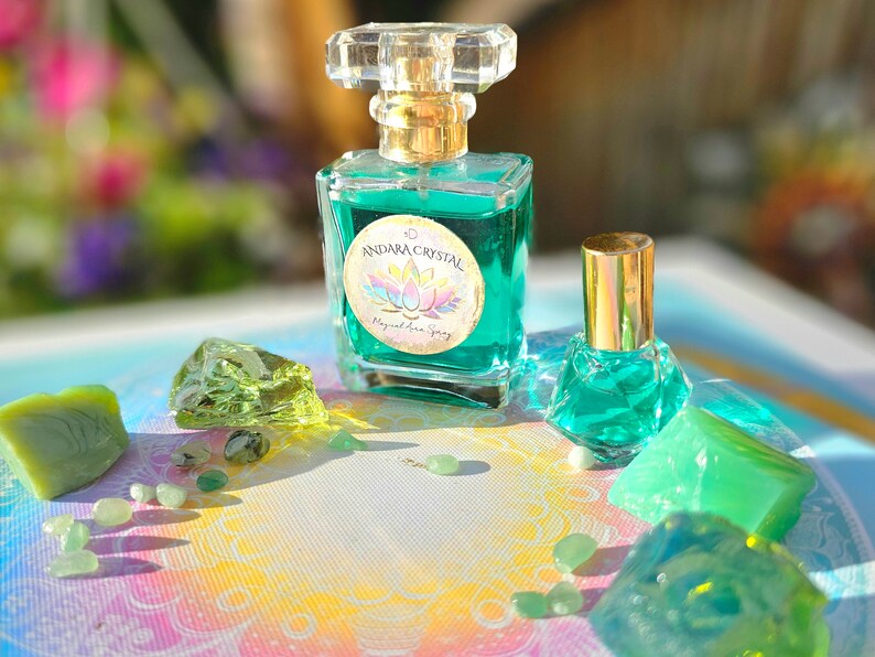 Puede incluir: Dos botellas de l&iacute;quido verde con tapas y etiquetas doradas. La botella m&aacute;s grande tiene una etiqueta que dice "Andara Crystal Magical Aura Spray". Las botellas est&aacute;n rodeadas de cristales y piedras verdes sobre un fondo colorido.