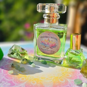 May include: A clear glass perfume bottle with a gold cap and a smaller matching bottle, both filled with green liquid. The label reads "Andara Crystal Magical Aura Spray". Green crystals and stones surround the bottles.