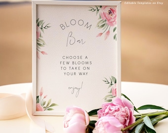 May include: A white framed sign with the text "BLOOM Bar CHOOSE A FEW BLOOMS TO TAKE ON YOUR WAY enjoy!"  The sign is decorated with watercolor pink flowers and green leaves. The sign is sitting on a beige table with a white ribbon and pink flowers.