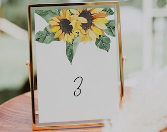 May include: A gold-framed table number card with a watercolor illustration of two sunflowers and green leaves. The number "3" is written in black cursive on the card.