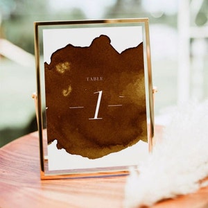 May include: A gold-framed table number card with a brown watercolor background. The card reads "Table 1".