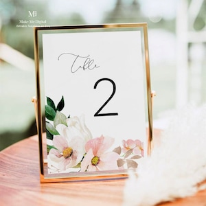 May include: A white table number card with a floral design and the text "Table 2" in black script font. The card is in a gold frame.