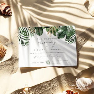 May include: A white wedding invitation with tropical leaf accents. The text reads "THE WEDDING CEREMONY of LILLIAN + THOMAS" with details about the ceremony's date, time, and location. The invitation is on a sandy surface with seashells.