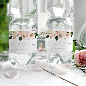 Könnte beinhalten: Zwei durchsichtige Plastikwasserflaschen mit weißen Etiketten, die mit einem Blumendesign und dem Text "Hannah & James 19.10.2022 | Kalifornien" verziert sind.