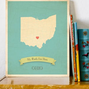 Könnte beinhalten: Ein blaugrüner Druck mit Vintage-Ästhetik, der eine Karte von Ohio mit einem roten Herzen in der Mitte zeigt. Der Text "My Roots Lie Here OHIO" ist unter der Karte gedruckt.