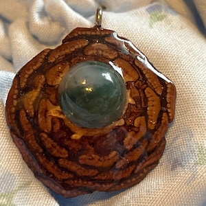 Peut inclure: Pendentif artisanal avec une base texturée marron foncé et une finition brillante. Une pierre sphérique verte est sertie au centre. Le pendentif est doté d'une bélière argentée pour la suspension. Le design a une esthétique naturelle et organique.