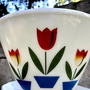 May include: A white milk glass mixing bowl featuring a repeating pattern of three tulips in red and yellow, each with green leaves and stems, planted in small blue pots. The bowl has a smooth, rounded rim and a tapered base design.