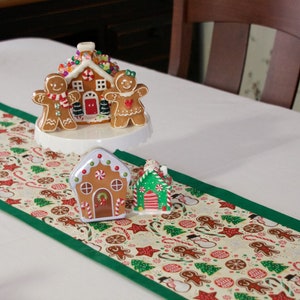 Puede incluir: Un camino de mesa blanco con un borde verde presenta una casa de jengibre y figuras de jengibre en un soporte para pastel blanco. El camino de mesa tiene un patrón repetitivo de figuras de jengibre, bastones de caramelo y árboles de Navidad sobre un fondo crema.