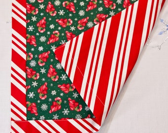 Op de afbeelding: Een kersttafelloper met een groen middenstuk met zuurstokken en sneeuwvlokken. De rand en de achterkant hebben rood-witte strepen. De loper is gemaakt van stof en is geschikt voor de feestdagen.