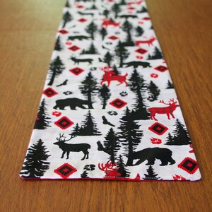 May include: A white table runner with a black and red woodland pattern. The pattern features silhouettes of deer, bears, and trees. The runner has a red border.