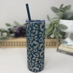 May include: A teal-colored stainless steel tumbler with a silver leopard print design. The tumbler includes a matching teal straw and lid. This insulated travel mug is designed to keep beverages cold or hot.