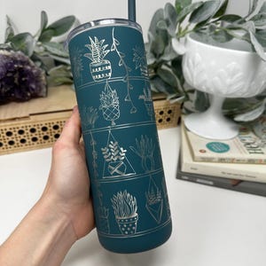 May include: Teal-colored stainless steel tumbler with a silver plant design. The tumbler features a lid and a matching teal straw. The design includes various potted plants and hanging plants. The tumbler is held by a person.