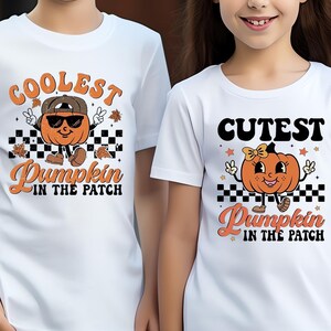 Può includere: Magliette bianche con grafiche di zucche. Una maglietta dice "COOLEST Pumpkin in the Patch" con una zucca con occhiali da sole e cappello. L'altra maglietta dice "CUTEST Pumpkin in the Patch" con una zucca con fiocco.
