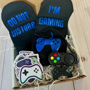 以下が含まれることがあります： 「DO NOT DISTURB」と「I'M GAMING」の青い文字がプリントされた黒い靴下のギフトボックス。青いコントローラーのグラフィック、ステッカー、キーホルダー、カラフルなボタンが付いた黒いポップイットのおもちゃも含まれています。ゲーマー向けの楽しいギフトです。