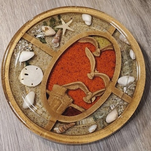 May include: Round resin art piece with a beach theme. The artwork features a central oval with a red background and carved birds. The outer ring is filled with sand, seashells, and small stones. The piece is encased in a clear resin.