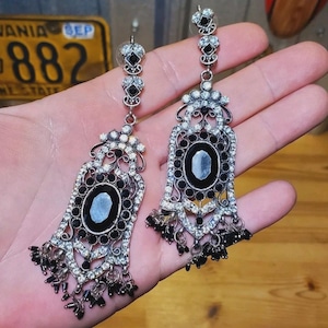 May include: A pair of ornate silver-toned earrings with black and clear crystal accents. Each earring features a large oval black stone, surrounded by smaller crystals and dangling black beads. The earrings are held in a hand.