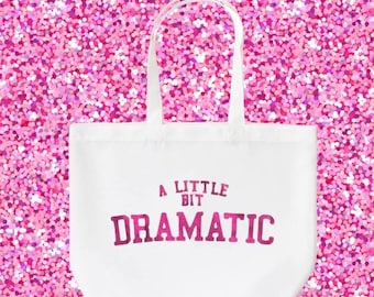Könnte beinhalten: Weiße Stofftasche mit rosa Schriftzug "A Little Bit Dramatic".
