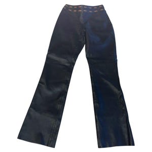 Puede incluir: Pantalones de cuero negro con diseño de pierna acampanada. Los pantalones tienen una cintura marrón con detalles rectangulares. El material es liso y brillante.