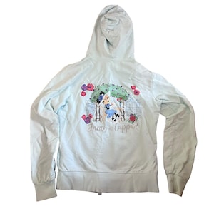 May include: Light blue zip-up hoodie with a hood and long sleeves. The back features a graphic of Minnie Mouse in a blue dress, seated at a table with the text "Fancy a Cuppa?" surrounded by flowers and a scenic background.