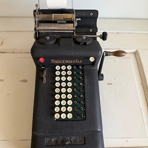 May include: A vintage black Burroughs adding machine featuring a manual hand crank with a wooden handle, a paper roll holder at the top, and a numeric keypad with white and black circular keys. The device includes a red non-add button and a total lever.