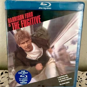 May include: A blue Blu-ray disc case for the movie The Fugitive starring Harrison Ford. The cover features a man running, with the text Harrison Ford is The Fugitive, and a circular blue sticker stating For use only with Blu-ray disc players and drives.
