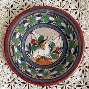 May include: A round ceramic plate featuring a hand-painted white rabbit in the center surrounded by green leaves and red floral accents. The plate has a wide dark red rim and a secondary inner border in dark gray with a repeating leaf pattern.