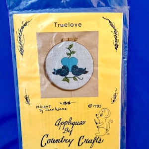 Op de afbeelding: Een knutselpakket in een doorzichtige plastic zak. De set bevat een cirkelvormig applicatieontwerp met twee blauwe vogels, een blauw hart en het woord "Truelove". De set is van Country Crafts, ontworpen door Joan Adams en gedateerd 1983.