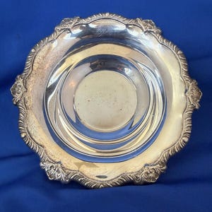 May include: A silver-colored, ornate dish with a scalloped edge and decorative floral accents. The dish has a circular, recessed center and a reflective surface. The background is a solid blue.