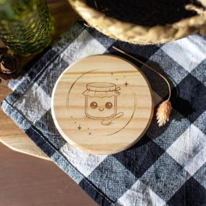 Peut inclure: Un dessous de verre rond en bois avec une illustration gravée d'un pot de miel souriant et d'une cuillère. Le dessous de verre est posé sur un tissu à carreaux noir et blanc.