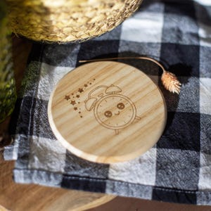 May include: A round, wooden coaster with a laser-etched design of a smiling alarm clock and stars. The coaster is light brown and sits on a black and white checkered cloth. A dried flower and woven basket are in the background.