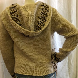 May include: A beige knitted hooded sweater with brown and white trim. The sweater has a loose fit and a long sleeve.