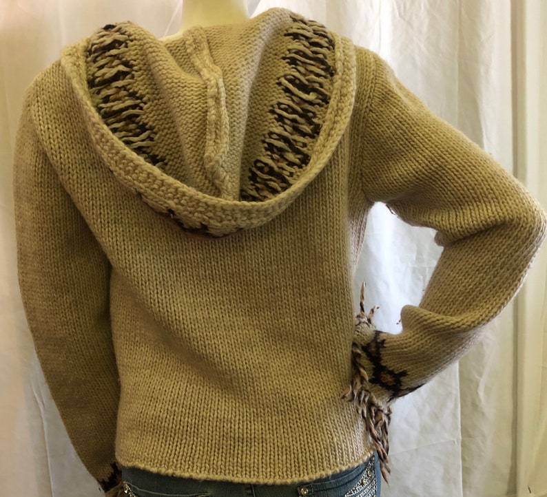 May include: A beige knitted hooded sweater with brown and white trim. The sweater has a loose fit and a long sleeve.