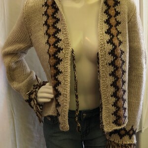 May include: A cream-colored knitted cardigan with brown and tan geometric patterns. The cardigan has a hood and a tie closure. The sleeves have fringe details.