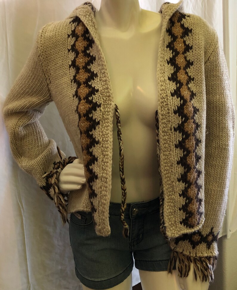 May include: A cream-colored knitted cardigan with brown and tan geometric patterns. The cardigan has a hood and a tie closure. The sleeves have fringe details.