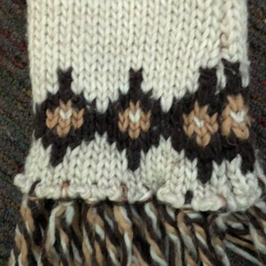 May include: A white knitted scarf with brown and tan geometric patterns and a fringe at the bottom.