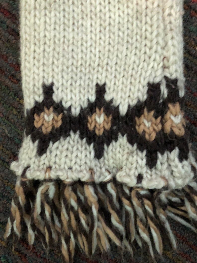 May include: A white knitted scarf with brown and tan geometric patterns and a fringe at the bottom.