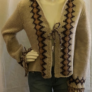 May include: A beige knitted cardigan with a brown and tan geometric pattern. The cardigan has a hood and a tie at the front. The sleeves have fringe at the cuffs.