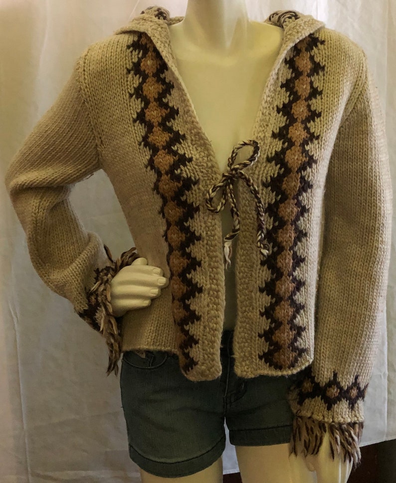 May include: A beige knitted cardigan with a brown and tan geometric pattern. The cardigan has a hood and a tie at the front. The sleeves have fringe at the cuffs.