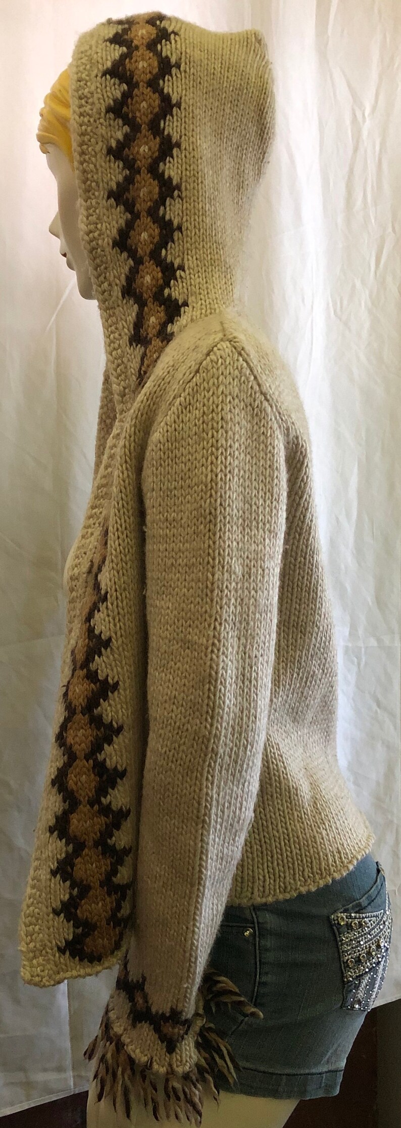 May include: A cream-colored knitted cardigan with brown and tan geometric patterns on the sleeves and hood. The cardigan has a fringed hem and is made of a soft, cozy material.