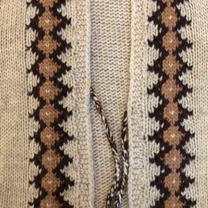 May include: Close-up of a knitted cardigan with a brown and beige geometric pattern on the front. The cardigan has a V-neck and a long, thin tie.
