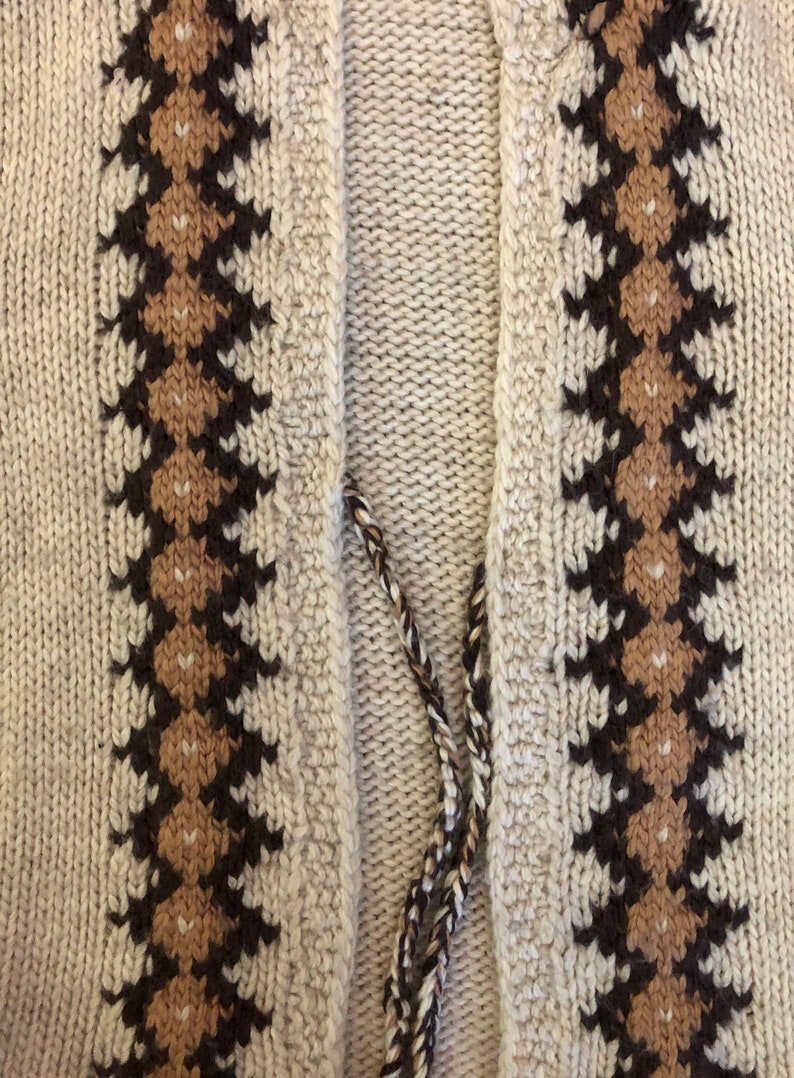 May include: Close-up of a knitted cardigan with a brown and beige geometric pattern on the front. The cardigan has a V-neck and a long, thin tie.