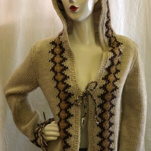May include: A beige knitted cardigan with brown and tan geometric patterns. The cardigan has a hood and a tie at the front. The sleeves have fringe at the cuffs.