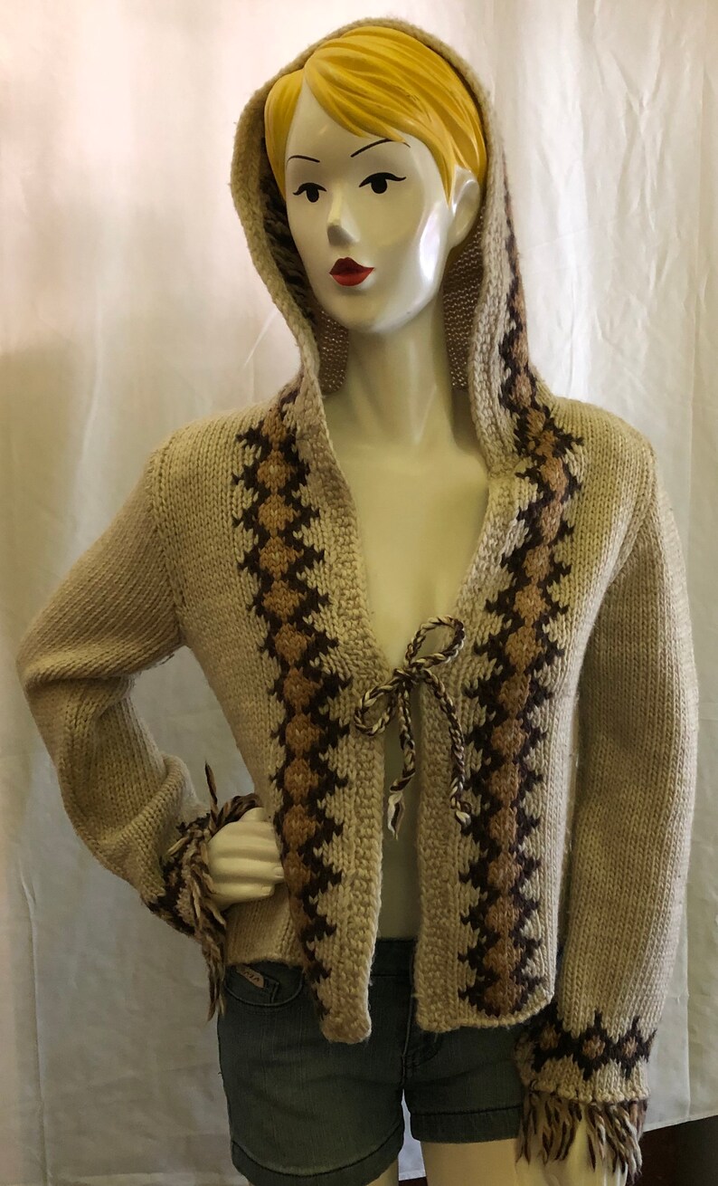 May include: A beige knitted cardigan with brown and tan geometric patterns. The cardigan has a hood and a tie at the front. The sleeves have fringe at the cuffs.