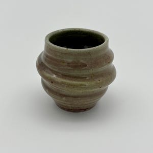 May include: A handmade ceramic cup with a unique, textured design. The cup features a spiral pattern around its body, with a blend of brown and green hues. The interior is dark, and the cup sits on a white surface.