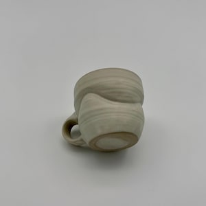 May include: A handmade, off-white ceramic mug with a unique, textured design. The mug features a rounded body and a small, curved handle. The exterior has a layered, organic appearance, suggesting a handcrafted process. The mug is suitable for coffee, tea, or other beverages.
