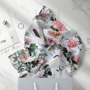 May include: A white silk scarf with a floral print featuring pink peonies, green leaves, and white birds. The scarf is folded and displayed on a white marble surface with a small gift bag and a business card that reads "creatsy" and "Created with Love!"