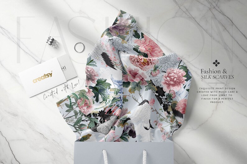 May include: A white silk scarf with a floral print featuring pink peonies, green leaves, and white birds. The scarf is folded and displayed on a white marble surface with a small gift bag and a business card that reads "creatsy" and "Created with Love!"