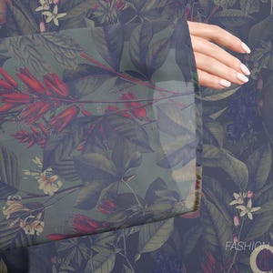 Puede incluir: Una tela verde y azul con un estampado floral que presenta flores rojas, hojas verdes y una flor blanca. La tela es transparente y tiene una mano que sale del lado.
