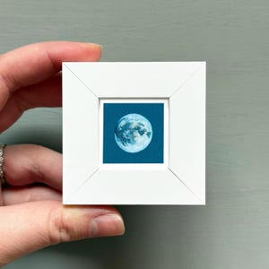 A hand is holding a small white picture frame containing a depiction of the moon.